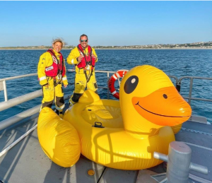 Two rescuers stand with a giant inflatable duck at the front of a boat.